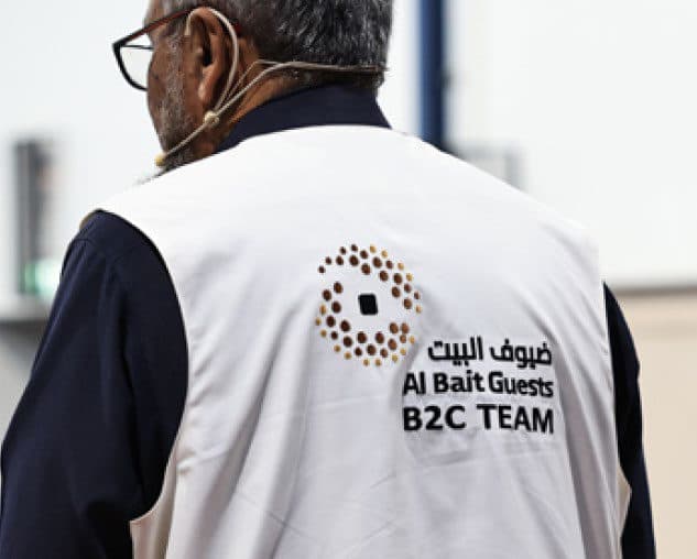أحد أفراد فريق ضيوف البيت يرتدي سترة بيضاء عليها شعار ضيوف البيت وعبارة B2C Team
