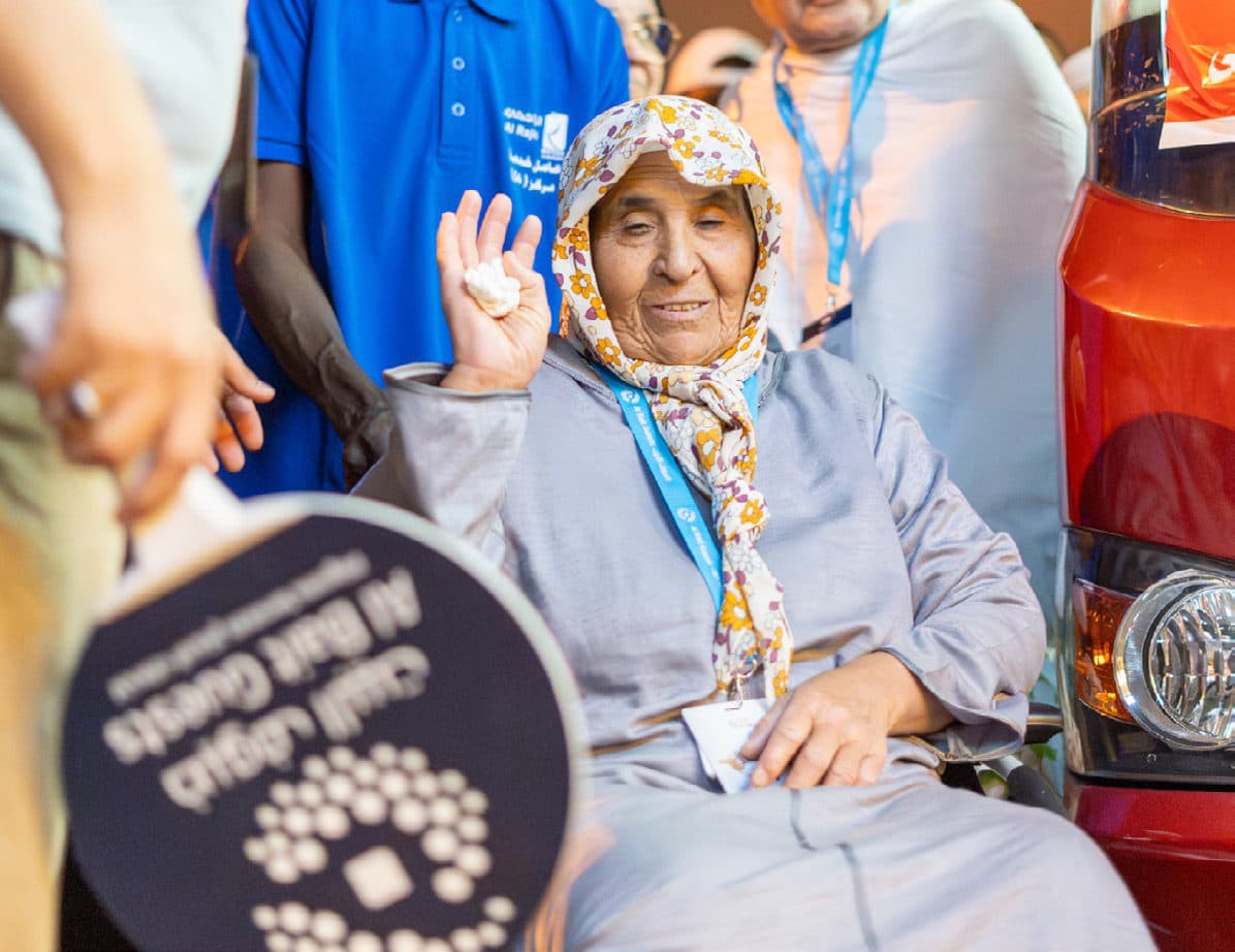 حاجّة مسنّة مبتسمة على كرسي متحرك تلوّح بيدها، ومن حولها موظفو ضيوف البيت