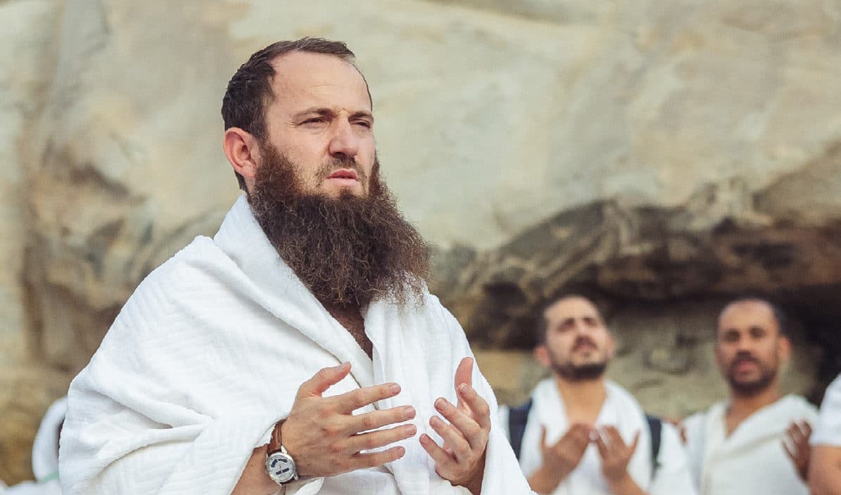 حاج بلباس الإحرام يرفع يديه بالدعاء، ومن خلفه حجاج آخرون